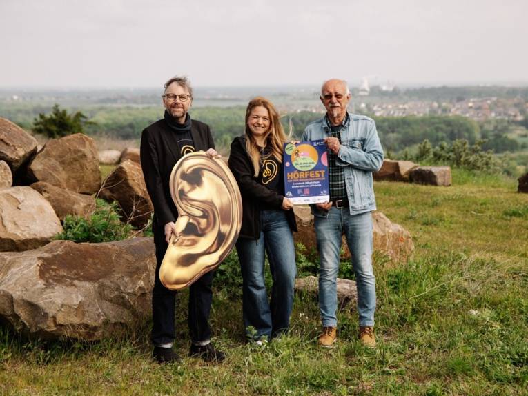 Zwei Männer und eine Frau stehen auf einer Wiese und halten ein Plakat und ein goldenes Ohr-Plakat in die Kamera. Im Hintergrund ist die Stadt und der Horizont zu sehen.