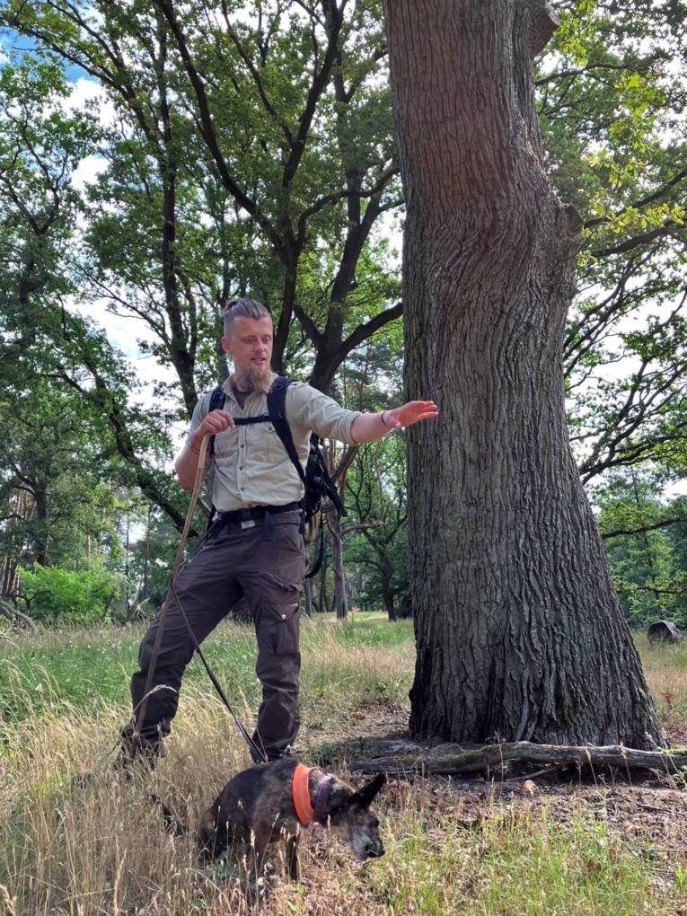 Ein Mann tr&auml;gt robuste Kleidung f&uuml;r drau&szlig;en und zeigt etwas in einem Gebiet mit Gr&auml;sern und B&auml;umen.