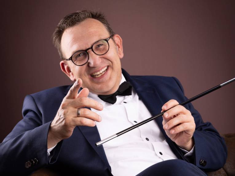 Lachender Mann mit kurzem Haar und Brille in Hemd, Fliege und Sakko hält einen Zauberstab.