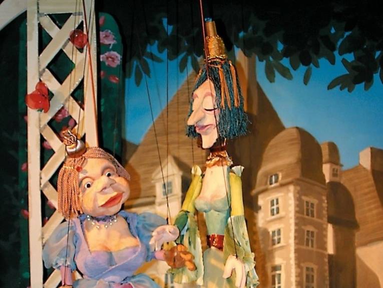 Zu sehen sind zwei Marionetten in Ballkleidern vor der Zeichnung eines Schlosses.