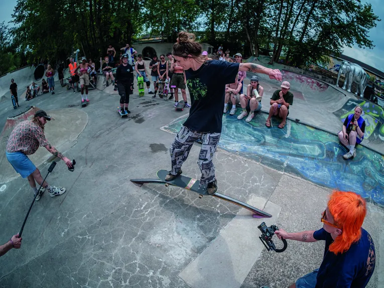 Eine dynamische Szene in einem Skatepark zeigt eine Person bei einem trickreichen Manöver auf einem Geländer. Rundherum verfolgen zahlreiche Zuschauerinnen und Zuschauer das Geschehen, einige mit Skateboards oder Kameras. Die weitwinklige Perspektive und die lebendige Atmosphäre vermitteln die Energie und Gemeinschaft einer Skateboard-Veranstaltung im Freien.