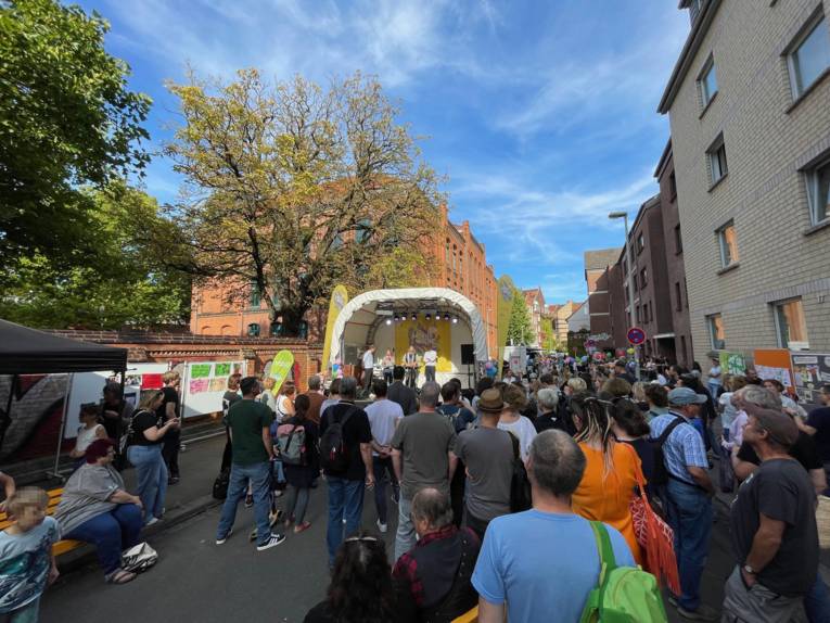 Eine Straße, auf der viele Menschen stehen aund auf eine Bühne sehen, rechts Wohnhäuser