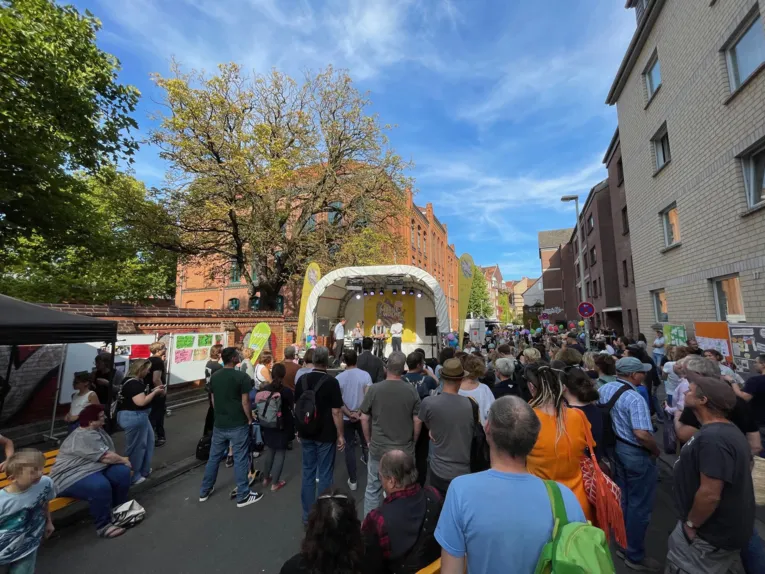 Eine Stra&szlig;e, auf der viele Menschen stehen aund auf eine B&uuml;hne sehen, rechts Wohnh&auml;user