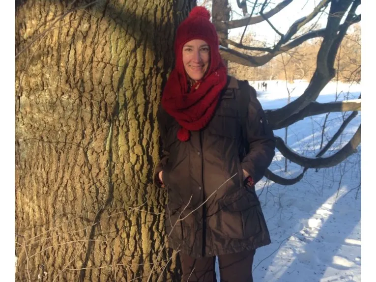 Eine Person mit Jacke, Schal und Müttze lehnt an einem Baum in winterlicher Umgebung.