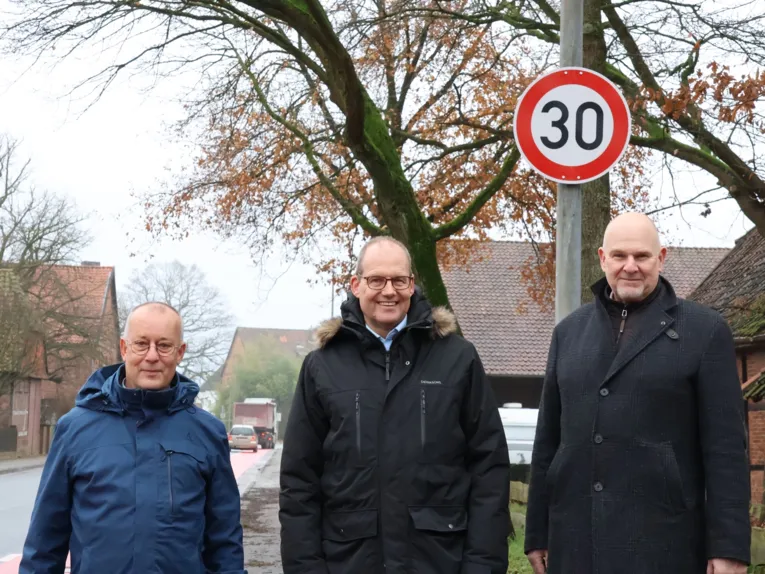 Drei Männer stehen auf dem Bürgersteig neben einer Straße, direkt hinter ihnen ist ein Schild "Tempo 30".