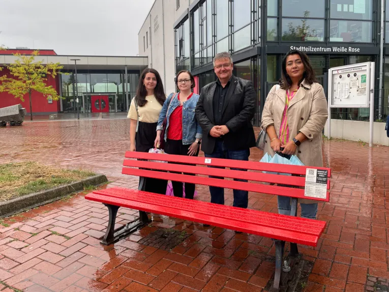 Vier Frauen und ein Mann stehen hinter einer rot angestrichenen Sitzbank auf einem Platz.