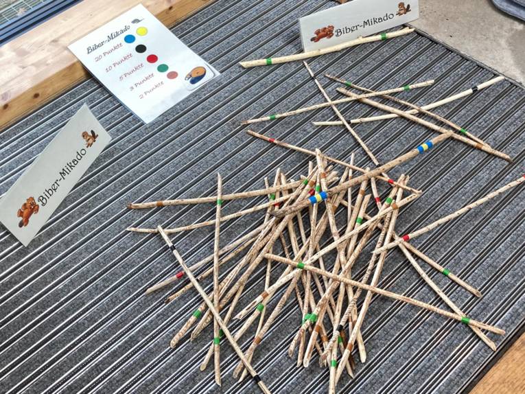 Holzst&ouml;ckchen unterschiedlicher Beschaffenheit sind jeweils farblich markiert und liegen in einem wilden Haufen aufeinander. Papieraufsteller tragen die Aufschrift "Biber-Mikado", eine Tafel liegt auf dem Boden zwischen den Papieraufstellern und gibt an, welche Farbe in diesem rustikalen Mikadospiel welchen Punktewert hat.