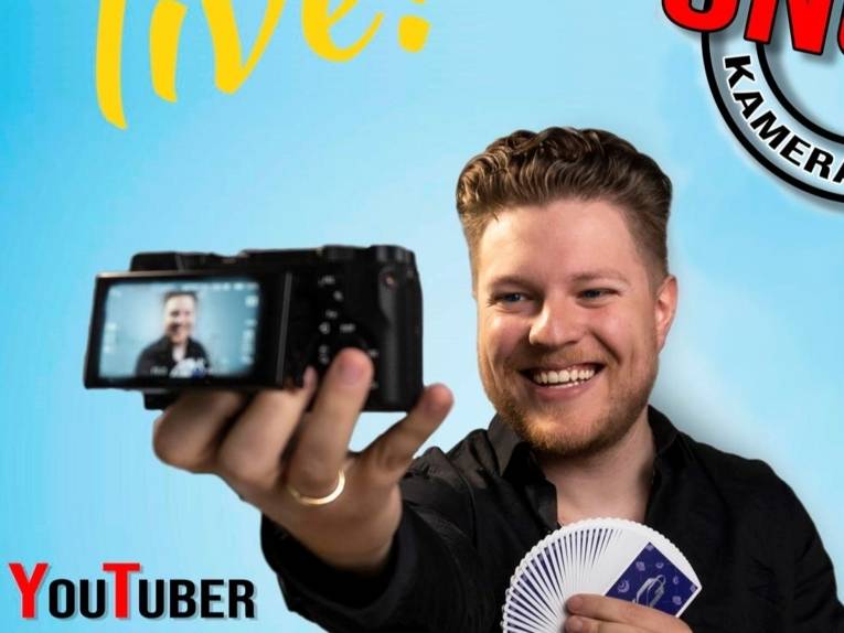 Zu sehen ist der der Youtuber Matthias Berger. Er hält aufgefächerte Spielkarten in einer Hand, während er mit der anderen Hand eine Videokamera auf sich selbst gerichtet hat.