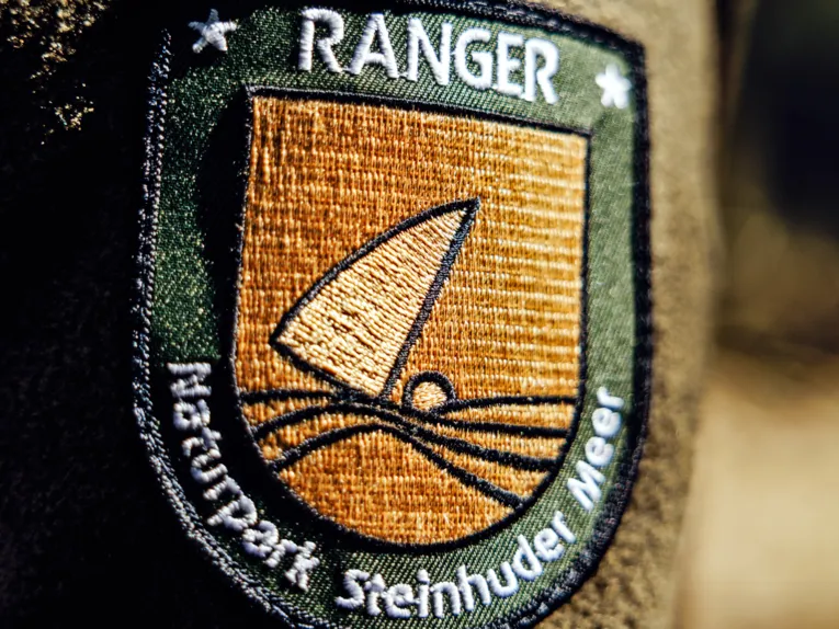 Aufnäher auf dem Ärmel einer Uniform eines Rangers im Naturpark Steinhuder Meer: "Ranger Naturpark Steinhuder Meer", dazu das Logo des Naturparks Steinhuder Meer, einem Segelboot mit Wasser und Land.