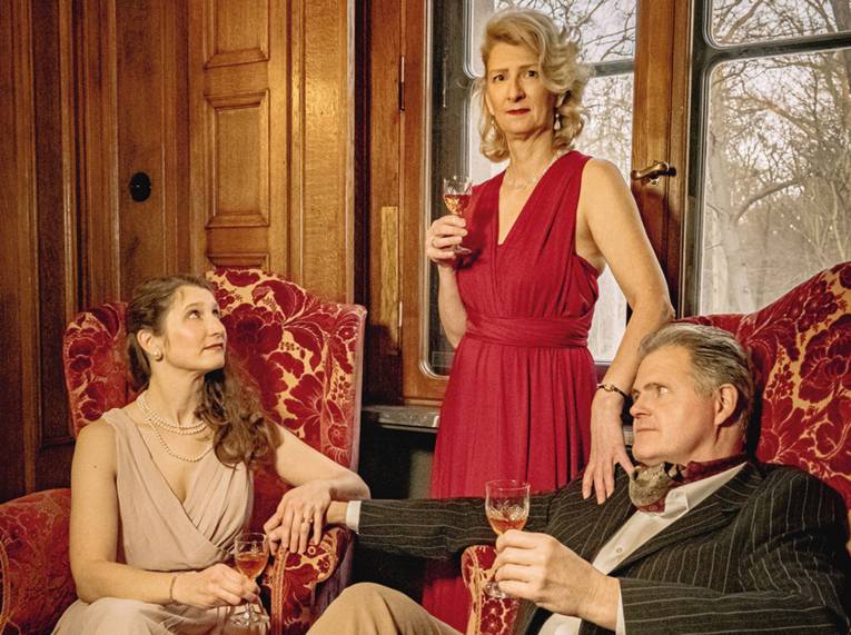 Zwei Frauen und ein Mann in eleganter Kleidung mit Gläsern in der Hand in einem Wohnzimmer