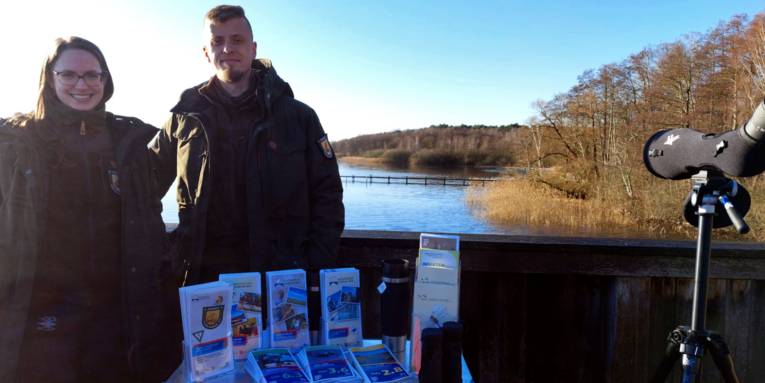 Zwei Personen tragen Kleidung f&uuml;r drau&szlig;en und stehen auf einer Aussichtsplattform. Auf einer Kiste vor ihnen ist Informationsmaterial. Im Hintergrund ist das Steinhuder Meer.