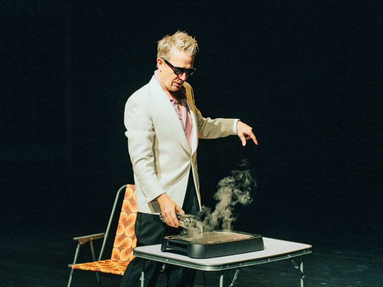 Ein Mann gießt eine Flüssigkeit auf einen Tischgrill, hinter ihm steht ein Campingstuhl.