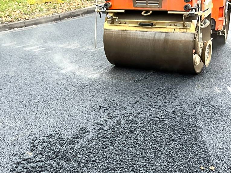 Auf einer dampfenden schwarzen Asphaltschickt rollt eine große Walze eines Fahrzeugs.