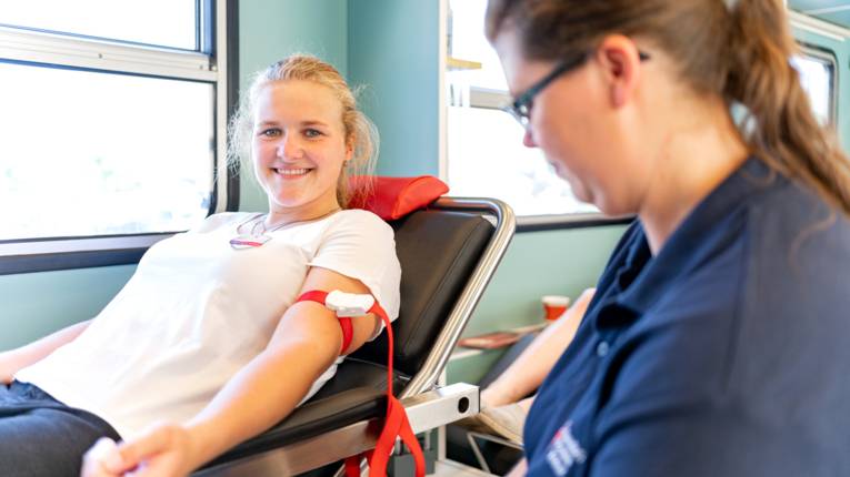 Eine Frau auf einer Liege beim Blutspenden. Davor sitzt eine eine Mitarbeiterin des Deutschen Roten Kreuzes.