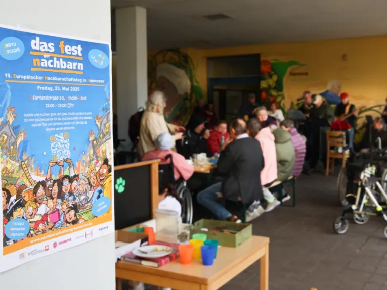 Vorn links ein blaues Plakat "das fest der nachbarn". Dahinter rechts ein Raum mit Menschen, die an einem gro&szlig;en Tisch sitzen.