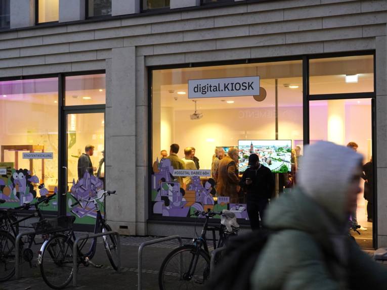 Blick von der Straße durch eine beleuchtete Fensterfront wie ein Schaufenster, dahinter stehen Menschen in einem Raum, ein großer Monitor ist zu sehen.