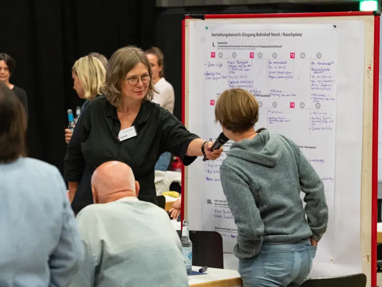 Vor einer Stellwand mit beschriebenem Papier h&auml;lt eine Frau einer anderen ein Mikrofon vor den Mund. Davor und dahinter sitzen und stehen Menschen.