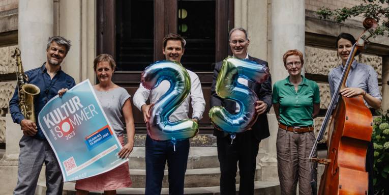 Sechs Personen stehen nebeneinander, Instrumente, eine 25 und ein Plakat zum Kultursommer 2023 der Region Hannover sind zu sehen.