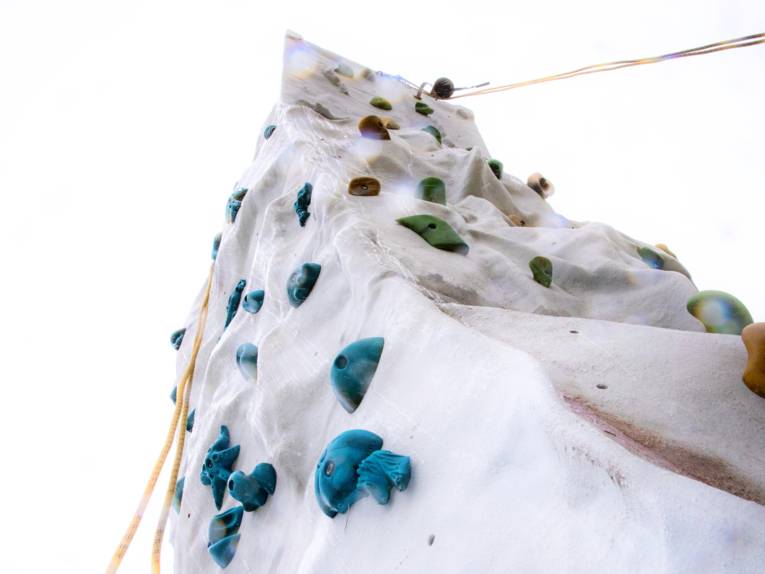 Blick von unten an eine freistehende Outdoor-Kletterwand mit bunten Klettergriffen und Sicherungsseilen.