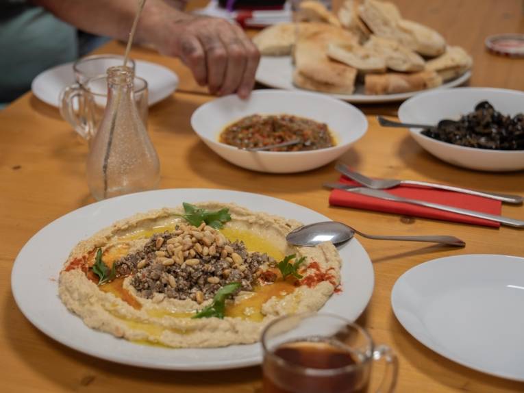 Ausschnitt eines Esstisches aus Holz, fotografiert von schräg oben. Auf dem Tisch mehrere Teller, gefüllt mit Hummus, Baba Ganoush und weiteren Speisen. Im Vordergrund ein halbgefülltes Teeglas.