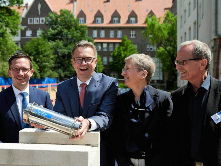 Drei Männer und eine Frau, einer der Männer hält einen zylinderförmigen Metallgegenstand in der Hand.