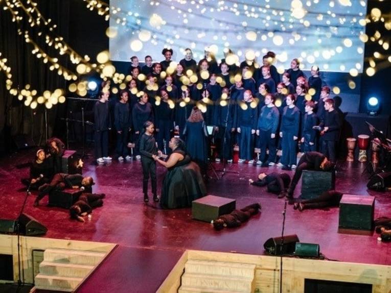 Zu sehen ist ein Chor und eine Performance mit in schwarz gekleideten Menschen. Lichterketten sind im Vordergrund zu sehen.