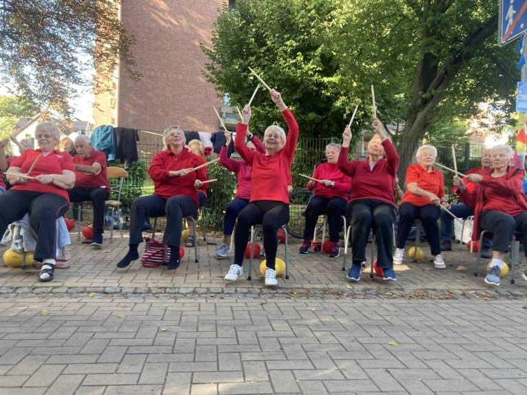 Alte Menschen sitzen auf St&uuml;hlen und strecken die Arme in die H&ouml;he mit St&ouml;ckern in der Hand. Sie lachen. Sie tragen alle rote T-Shirts