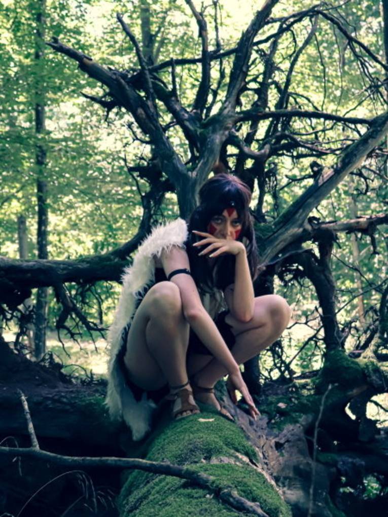 Eine Frau hockt verkleidet auf einem Baumstamm in einem Wald.