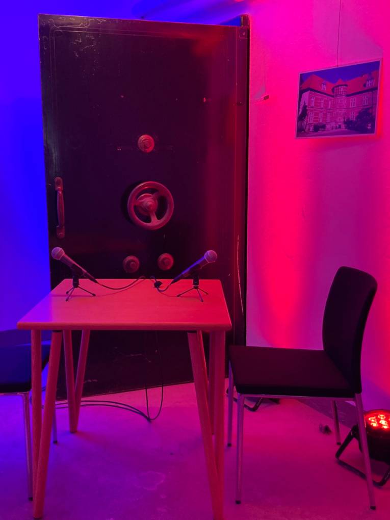 Neben der schweren Panzertür eines Tresors steht ein Tisch, daran zwei Stühle und darauf zwei Mikrofone. Scheinwerfer tauchen die Szene in Rot- und Blautöne.