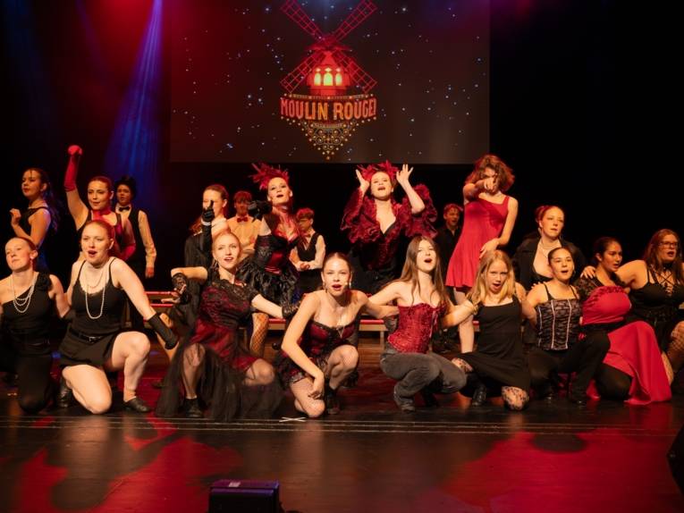 Zu sehen ist eine Gruppe auf der Bühne, gekleidet im Stile von Moulin Rouge.