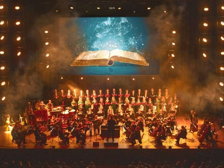 Zu sehen ist ein Chor und ein Orchester auf einer Bühne. Im Hintergrund ist eine große Leinwand zu sehen, auf der aus einem geöffneten Buch Funken und Nebelschwaden ausströmen.