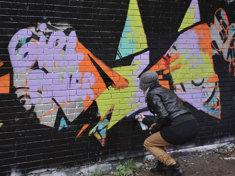 Eine schwarze Mauer. Davor steht eine Person und sprayt bunte Elemente darauf.