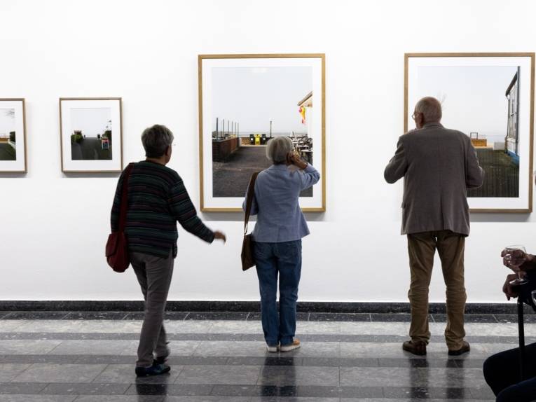Besucherinnen und Besucher schauen sich Bilder an, die nebeneinander an einer Wand ausgestellt sind.