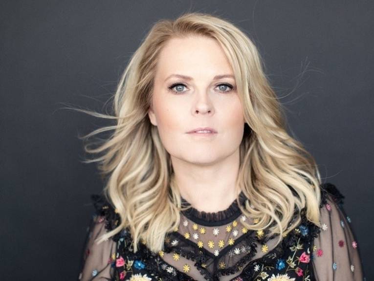 Eine blonde Frau mit einem schwarz geblümten Oberteil schaut ernst in die Kamera.