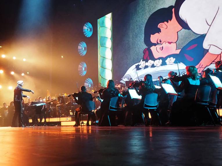 Ein Orchester spielt vor einer Leinwand, auf der ein Ausschnitt des Disney-Films „Schneewitchen und die 7 Zwerge“ zu sehen ist