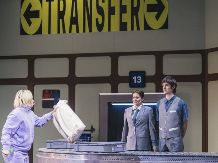 Check-In-Szene auf einer Bühne: Eine Frau legt ein Gepäckstück in einen Behälter auf einem Rollband, hinter dem eine Frau und ein Band in Uniform eines Flughafensicherheitsdienstes stehen.