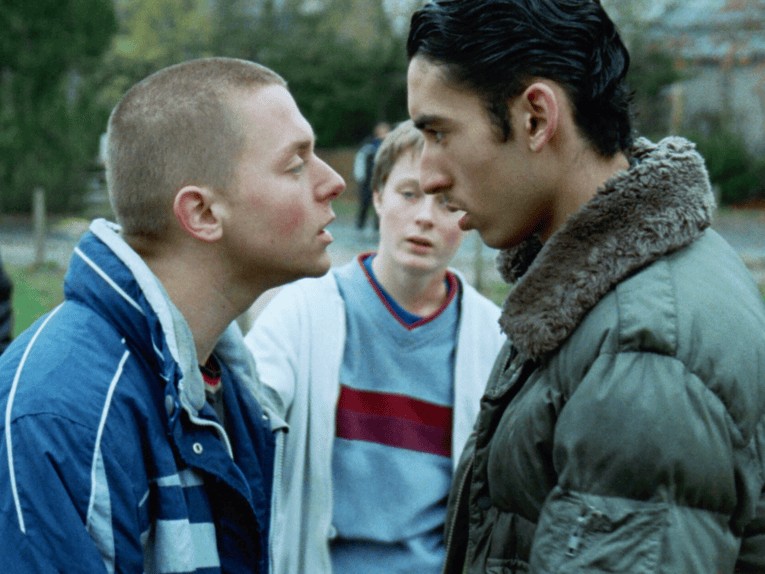 Zwei junge Männer stehen sich gegenüber und gucken sich feindselig an. Im Hintergrund steht ein weiterer junger Mann und guckt zu.