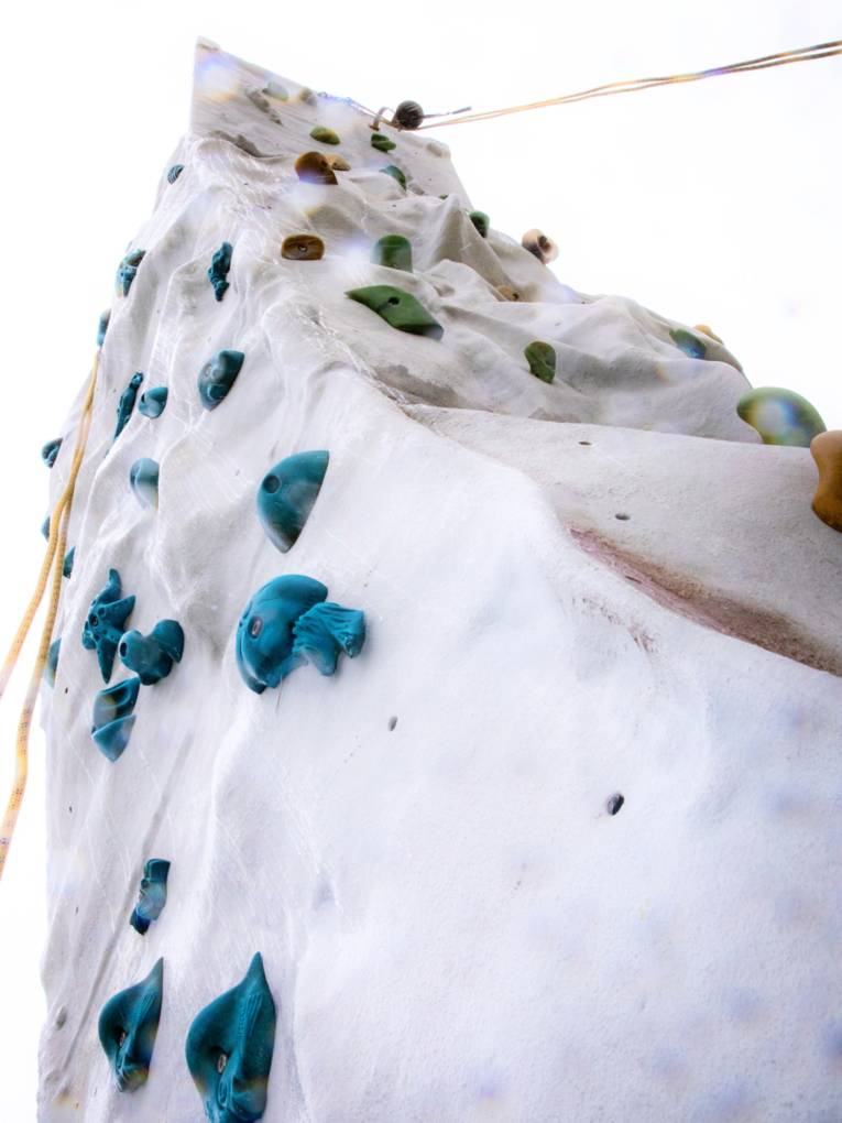 Blick von unten an eine freistehende Outdoor-Kletterwand mit bunten Klettergriffen und Sicherungsseilen.