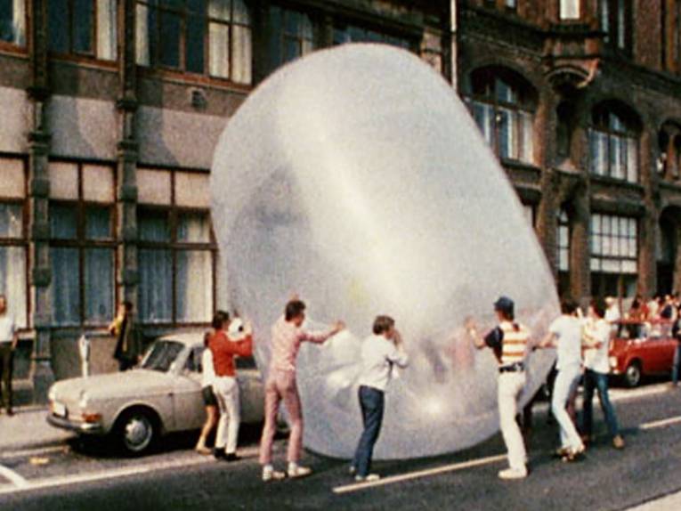 Mehrere Personen rollen einen riesigen Gummiball eine Straße entlang