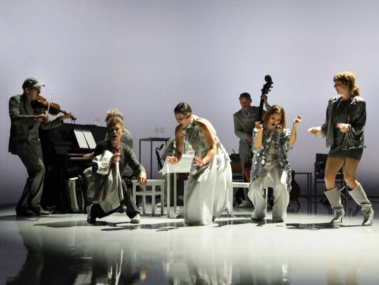 Auf einer Bühne im Hintergrund ein Violinist, ein Klavierspieler, ein Saxofonist und ein Bassist, davor singen tanzend drei Frauen und ein Mann.