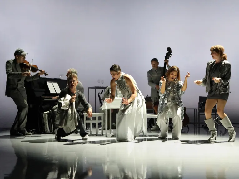 Auf einer Bühne im Hintergrund ein Violinist, ein Klavierspieler, ein Saxofonist und ein Bassist, davor singen tanzend drei Frauen und ein Mann.
