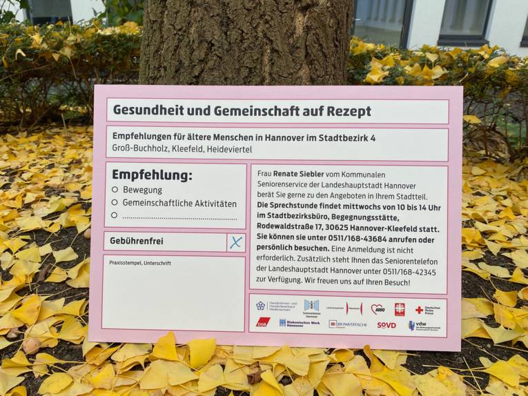 Ein überdimensionales ärztliches Rezept steht an einen Baum gelehnt, drum herum Herbstlaub