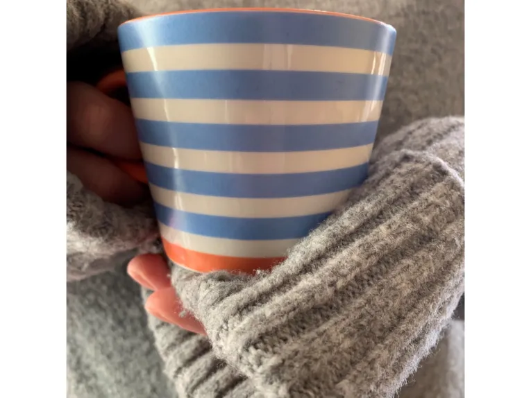 Hände in einem dicken Strickpulli, die eine blau-weiß gestreifte Tasse halten.