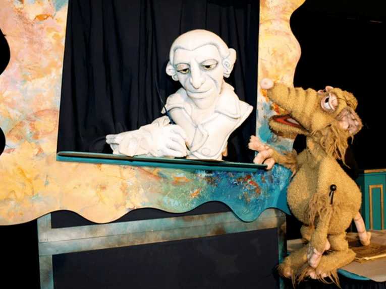 Zu sehen ist das Bühnenbild des Stücks. In einem leeren Bilderrahmen steht die Figur des Haydn – eine sprechende Büste. Daneben ist die Puppe einer großen Ratte zu sehen, die scheinbar zur Büste spricht.
