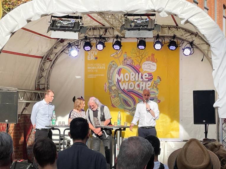 Eine Bühne, auf der drei Männer und eine Frau stehen, hinter ihnen ein Plakat  mit der Aufschrift Mobile Woche.