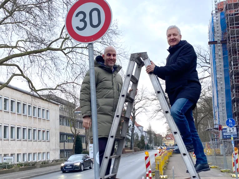 Zwei Männer stehen auf einer Leiter an einer Straße an einem Verkehrsschild mit einer rot umrandeten Zahl 30.