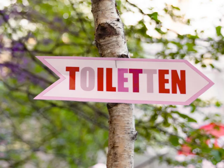 Ein Wegweiser aus Holz ist an einen Baum getackert, darauf steht "Toiletten".