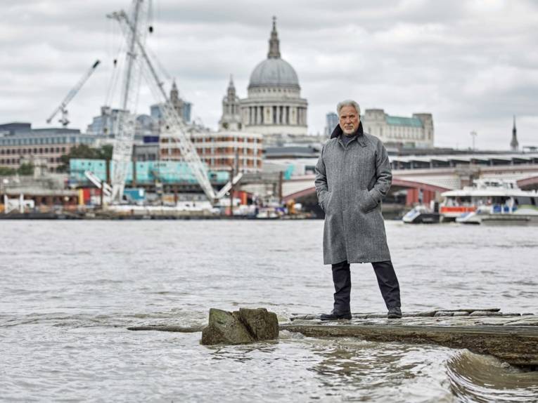Ein Mann in grauem Mantel steht am Wasser, im Hintergrund ist ein Hafen sowie Gebäude zu sehen.