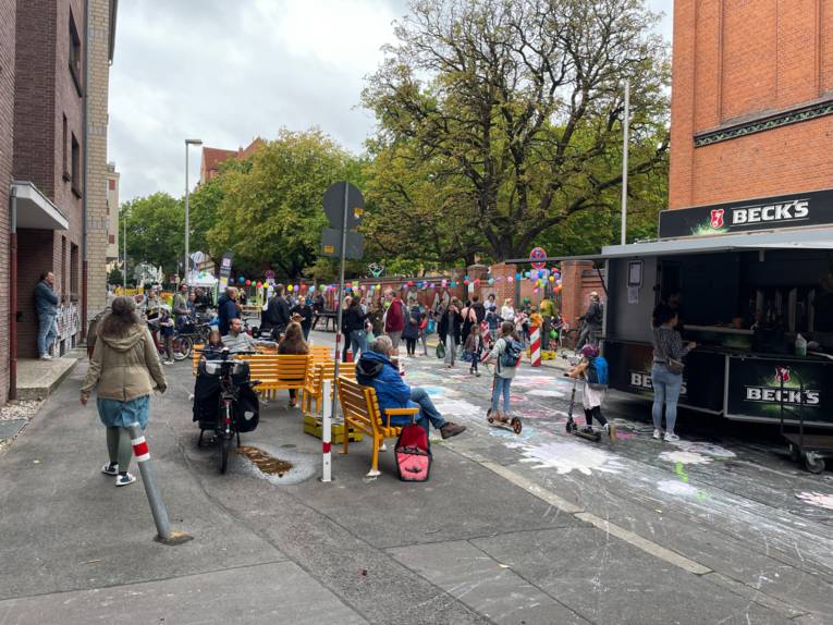 Eine geschmückte Straße mit Menschen, rechts ein Getränkewagen. Die Menschen gehen auf der Straße, die mit Kreidebildern von Blumen bemalt ist, rechts eine lange Girlande aus Luftballons, gelbe Bänke stehen auf der Straße, auf der Menschen sitzen.