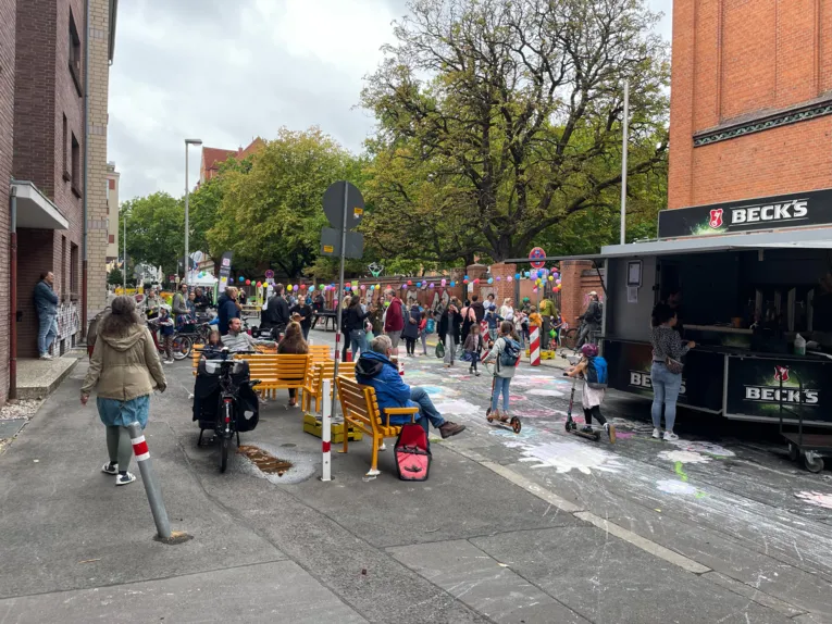 Eine geschm&uuml;ckte Stra&szlig;e mit Menschen, rechts ein Getr&auml;nkewagen. Die Menschen gehen auf der Stra&szlig;e, die mit Kreidebildern von Blumen bemalt ist, rechts eine lange Girlande aus Luftballons, gelbe B&auml;nke stehen auf der Stra&szlig;e, auf der Menschen sitzen.